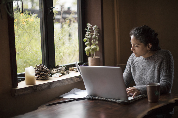 Perfect set up to work at home: natural light filters through the window illuminating one young woman of mixed-race who leans into a redwood-slab table top desk while typing on her laptop and reading reference papers. A houseplant, collected pine cones, and a wax candle sit on the wooden window sill. She has a mug of tea and is wearing a cozy gray wool sweater. This could be a back-to-school scene or a working-on-deadline from home scene. Themes of indoors/outdoors and work/life balance are apparent.
