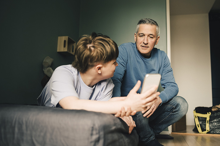 Mature man and son discussing over smart phone in bedroom at home